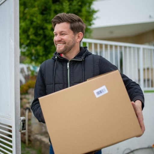 A happy man with a beard carries a large box outside a modern house.